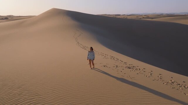 A young women with long hair wearing a summer dress and walking barefoot across vast sand dunes