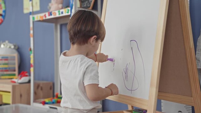 Boy drawing on easel whiteboard with right hand in studio among toys and storage shelves, marker scribbles visible; curiosity creative play.