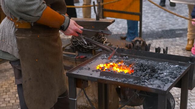 Blacksmith forging metal at an outdoor forge. Skilled blacksmith wearing a leather apron using tongs to heat a metal object in hot glowing coals at a traditional outdoor forge