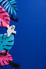 Naklejka premium Tropical Halloween vertical paper ghost and bats among colorful monstera leaf on blue background