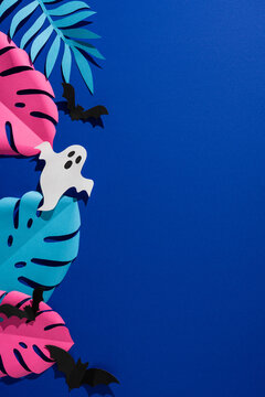 Tropical Halloween vertical paper ghost and bats among colorful monstera leaf on blue background