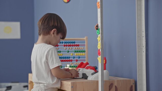 Boy playing with toy cash register and wooden abacus, hands pressing buttons at a table inside a building; curiosity learning.