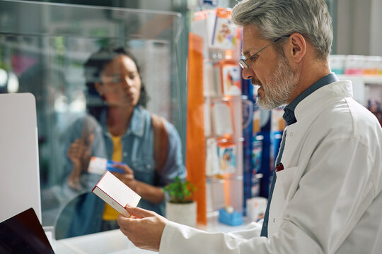 Mature pharmacist holding box of medicine while talking to customer in pharmacy,