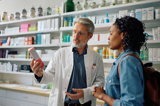 Mature pharmacist advising black woman in choosing skincare product in pharmacy.