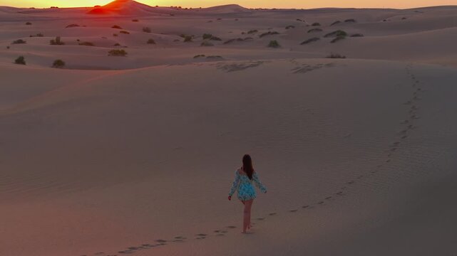 A young women with long hair wearing a summer dress and walking barefoot across vast sand dunes