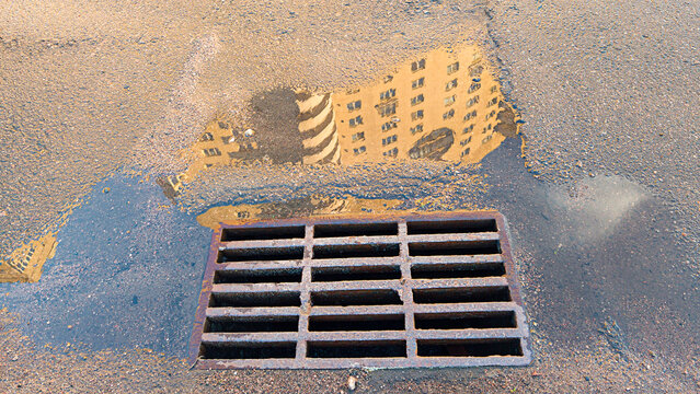 Urban building reflecting in a water puddle on pavement, showing worn metal storm drain next to infrastructure element