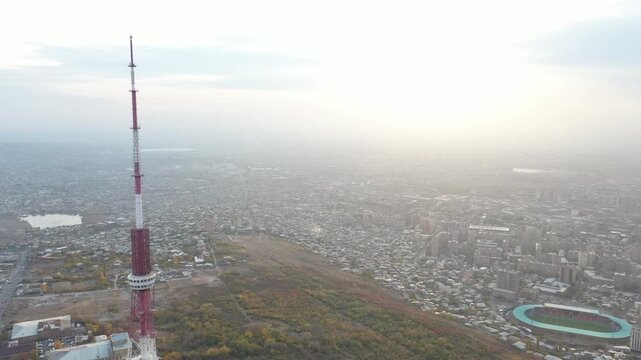 An aerial drone view of the Yerevan TV Tower standing tall over the misty Armenian capital during autumn with colorful forest and scenic city panorama in the background.

