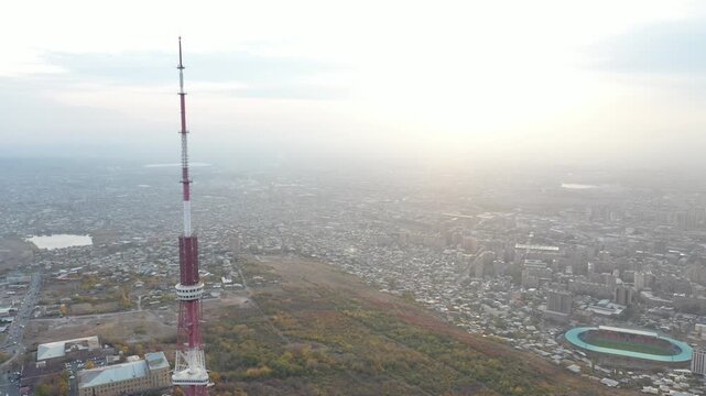An aerial drone view of the Yerevan TV Tower standing tall over the misty Armenian capital during autumn with colorful forest and scenic city panorama in the background.

