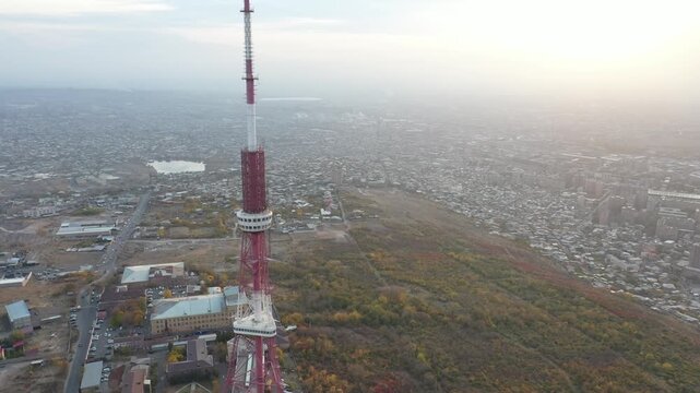 An aerial drone view of the Yerevan TV Tower standing tall over the misty Armenian capital during autumn with colorful forest and scenic city panorama in the background.

