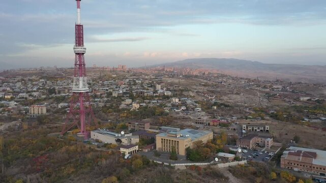 An aerial drone view of the Yerevan TV Tower standing tall over the misty Armenian capital during autumn with colorful forest and scenic city panorama in the background.

