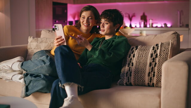 Laughing women holding smartphone sitting cozy living room. Smiling friends 