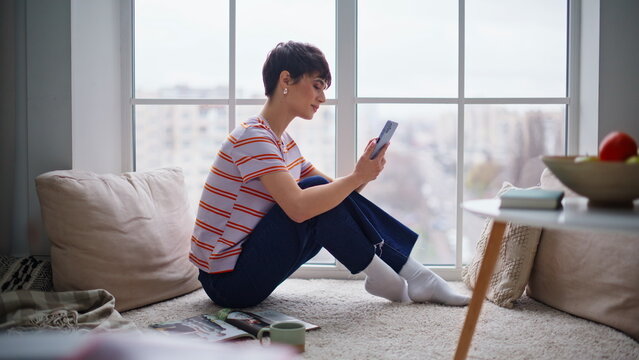 Cheerful lady reading smartphone message sitting home floor at domestic weekend.