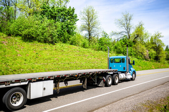 Blue day cab big rig semi truck with empty long flat bed semi trailer running on the narrow green road