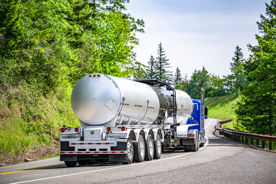 Bright blue big rig semi truck transporting liquid cargo in long tank semi trailer with four axels running on the winding road with hills and forest on the sides