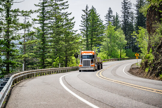 Classic orange big rig semi truck tractor transporting fastened cargo on flat bed semi trailer driving on the winding mountain road with forest and rock cliff