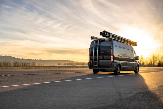 Commercial dark gray cargo mini van with ladder on the backside and pipes on the roof running on the road at sunset time