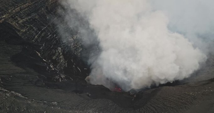 Mount Yasur crater belching thick white and gray smoke while glowing magma lights the vent, dramatic volcanic power on Tanna Island, Vanuatu, raw and explosive landscape. Aerial view drone shot