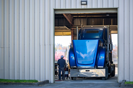 Mechanic performs maintenance and routine repairs on the blue big rig semi truck tractor with open hood standing in an auto repair shop