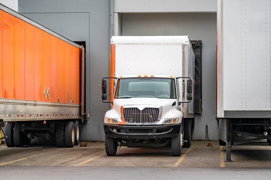 Middle duty white day cab rig semi truck with box trailer loading cargo standing at warehouse dock gate beside another semi trailers