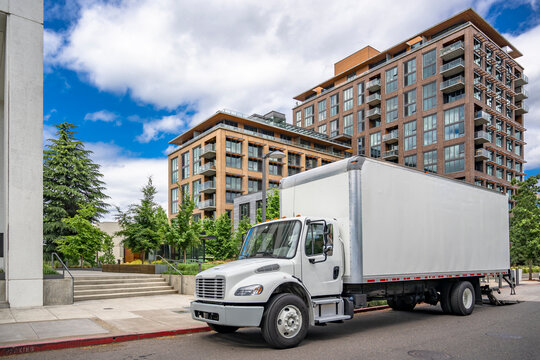 Middle duty white day cab rig semi truck with long box trailer unloading cargo standing on the urban city street with multilevel apartment and with lift platform lowered at the rear of the trailer