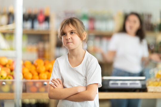 Angry child is standing in grocery store, capricious and sulky silence. Girl makes faces and ignores conversation with her mother, parent screams and is indignant in background