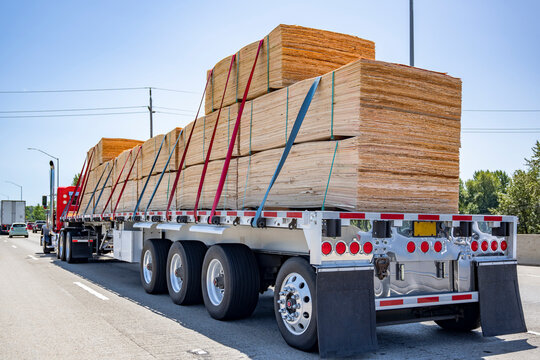 Red classic big rig semi truck transporting fastened lumber cargo on the two flat bed semi trailers driving on the wide highway road
