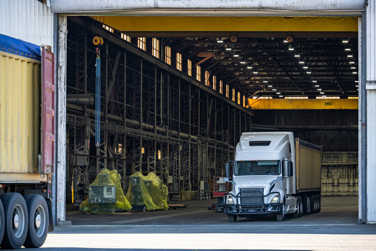 White big rig semi truck with container semi trailer loading cargo standing inside warehouse building