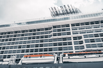 Low angle view of a large white cruise liner with rows of cabin windows, orange lifeboats and upper deck funnels against a cloudy sky for travel and maritime themes © skyNext