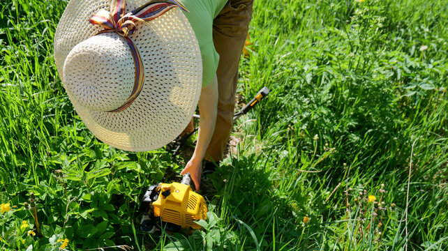 Woman in sun hat using string trimmer to cut tall grass in outdoor rural field during gardening and yard maintenance work