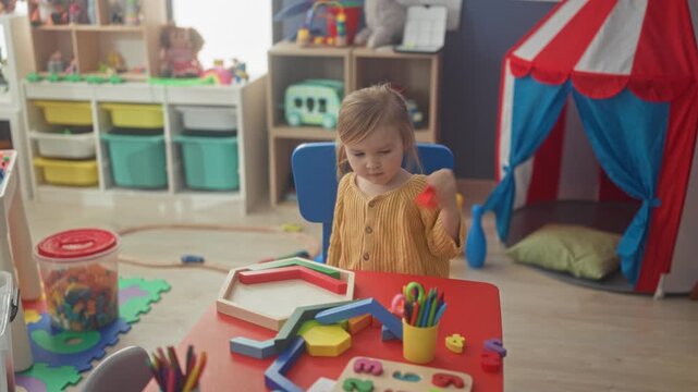 Girl placing a wooden block with her hand on a red table among puzzle pieces, crayons and toys in a playroom; quiet concentration and focused play.
