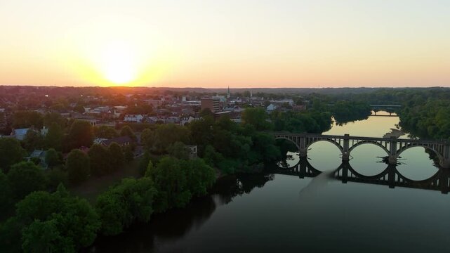 Fredericksburg, Virginia, scenic 4K aerial video establishing the historic downtown district and a concrete arch bridge over the calm Rappahannock River during a vibrant golden hour sunset.