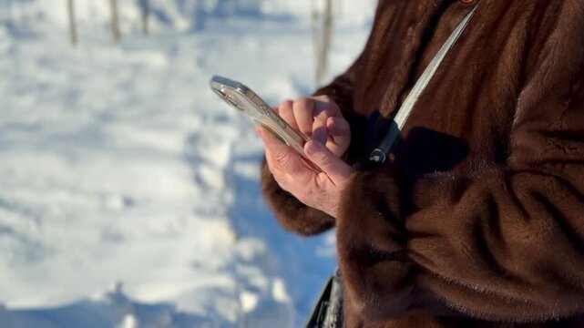 Woman texting on smartphone in snow luxury mink coat closeup of hands with manicure sunlight glinting on icy landscape urban