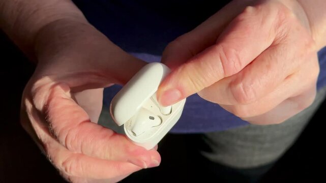 Closeup woman holding white earbud case, gently removing single earbud for listening warm sunlight highlights textured hands