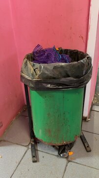 Close-up green trash can overflowing with purple cloth and scrubber, plastic liner packed with food scraps and crumpled packaging