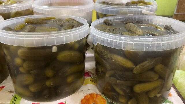 Close-up pickles plastic buckets market vendor display, brine filled cucumbers packed in clear containers on colorful tablecloth