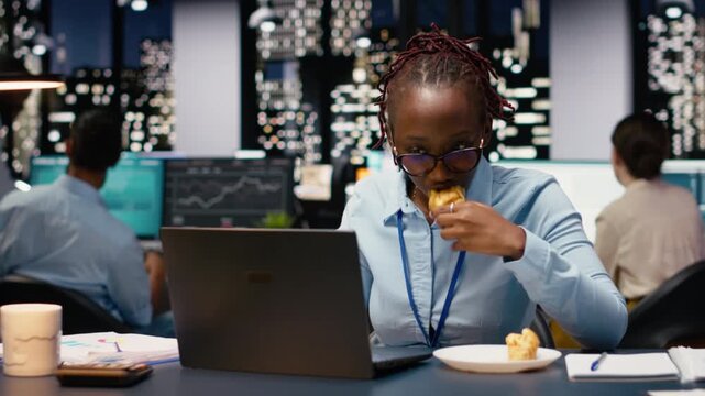Stressed businesswoman dealing with eating disorder during late night work session. Young woman eats excessive junk food and carbs at her desk while reviewing data on computer, temptation.