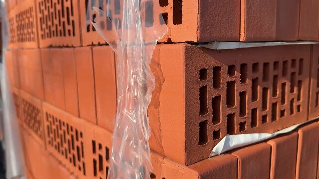 Close-up red bricks stacked outdoors, plastic wrap glistening in sunlight, perforated hollow bricks on pallet at hardware store