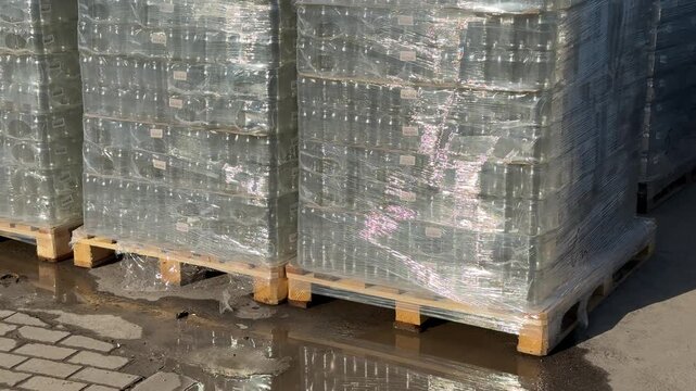 Pallets of glass jars wrapped outdoors, wooden pallets lined on sidewalk outside hardware store, shrink-wrap glinting in sunlight