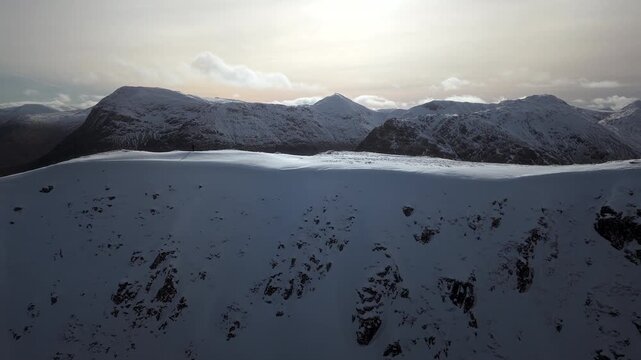 Lateral aerial drone shot of a distant hiker on snow covered mountains near Glencoe, Scotland under cloudy winter sky.
