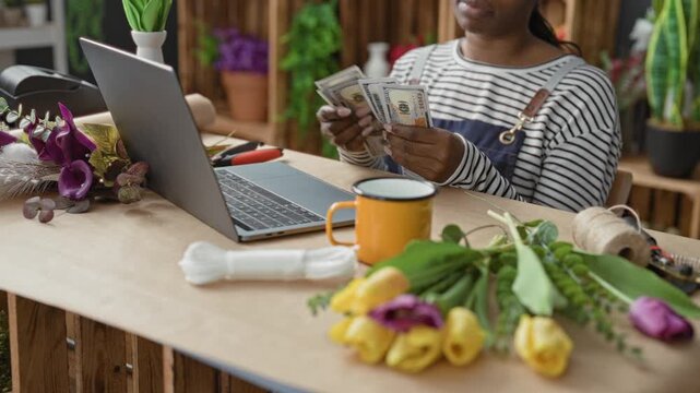 Woman florist counting dollars with hands and typing on laptop among tulips and spool at studio; focused small business.