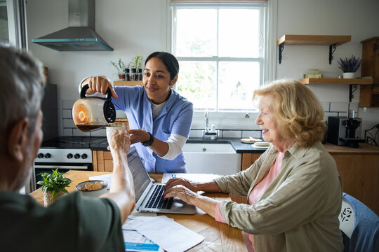 Caregiver pouring coffee as seniors use laptop in home kitchen
