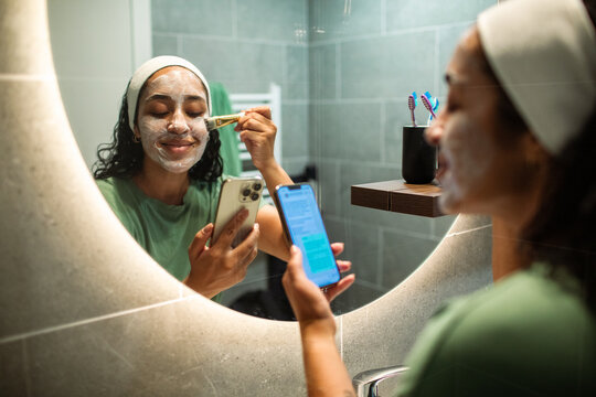 Young woman applying face mask while texting in home bathroom mirror