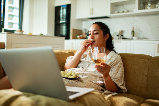 Woman relaxing on sofa with wine and grapes, streaming on laptop at home