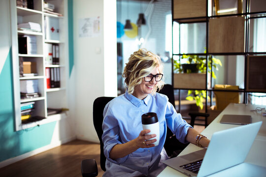 Smiling woman with coffee working on laptop in modern office