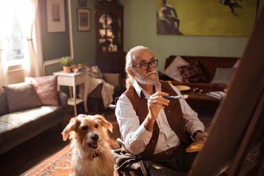 Senior man in wheelchair painting on canvas at home with dog