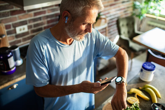 Active mature man checking smartwatch while prepping smoothie in home kitchen