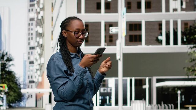 African american woman walking on city street using smartphone, relaxing by looking at social media posts on feed. Person downtown metropolitan avenue scrolling phone on sidewalk.