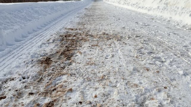 Snow cleared pedestrian path alongside highway, showing salted and grit strewn surface, high plow berm, tire tracks and compacted