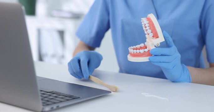 Dentist in medical gloves showing how to clean teeth with model of jaw during online consultation by laptop at white table in clinic, closeup