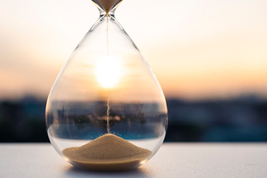 Close up of an hourglass with golden sand flowing against a bright sunset sky background. Concept of time passing, business deadline, future planning, life countdown, and hope.